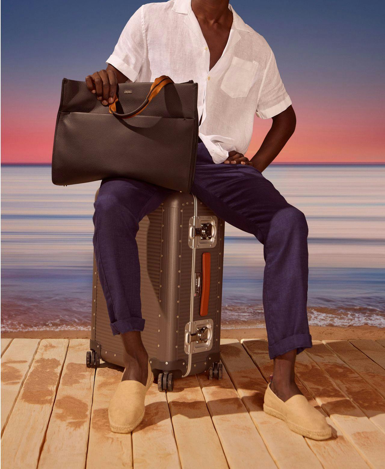 male model sitting infront of ocean on luggage wearing chinos and sport shirt holding a tote bag