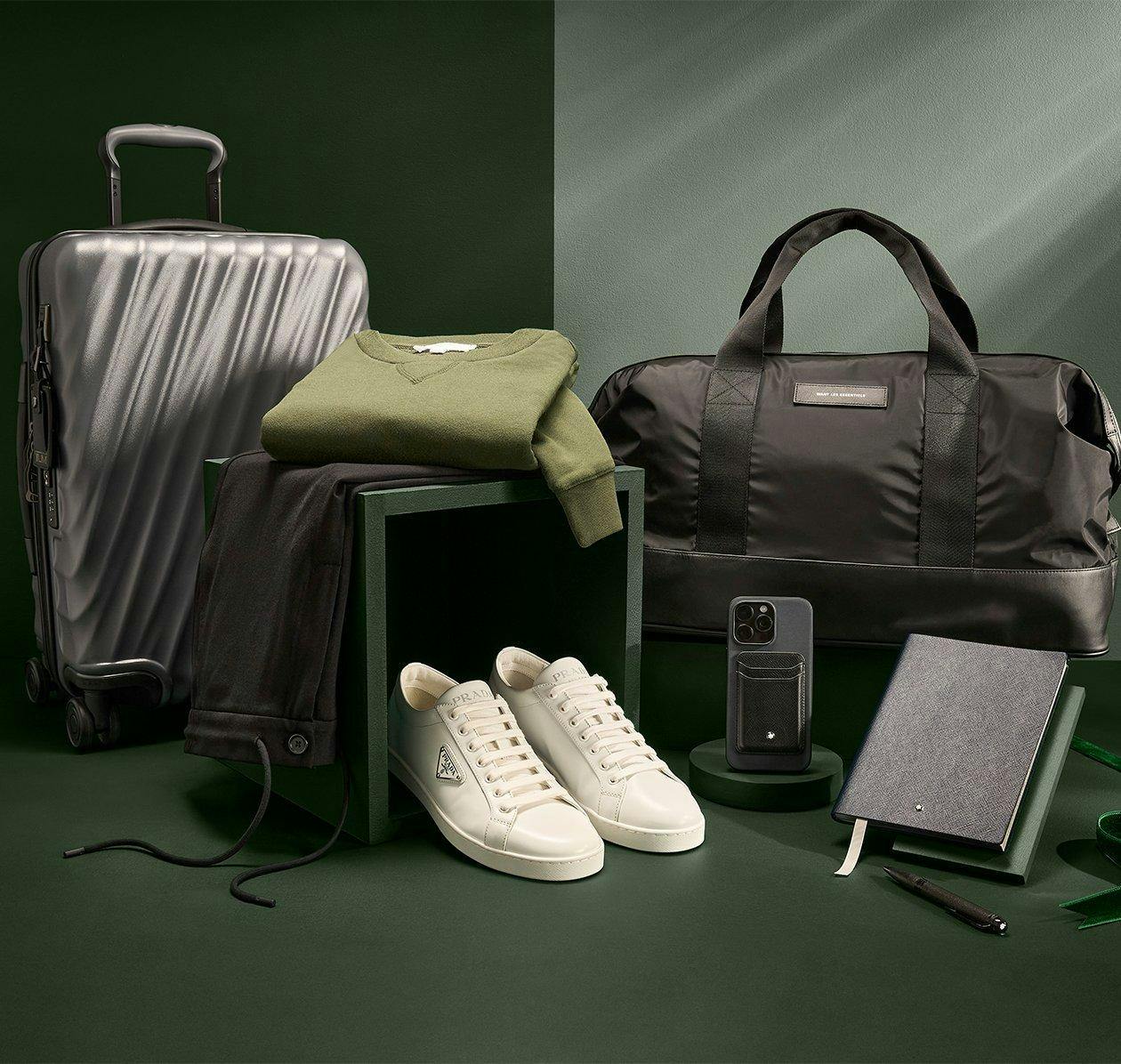Prada white sneakers, iPhone case, grey notebook and pen, green sweater, Tumi suitcase, and black pants
