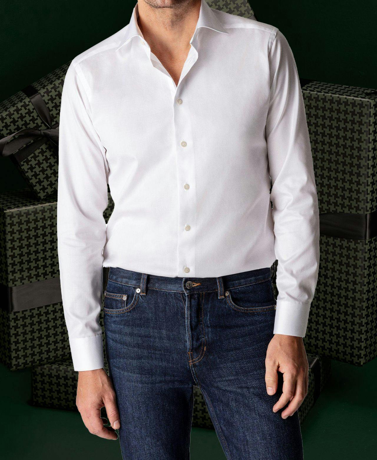 Male model showcases white shirt and blue jeans against dark green background