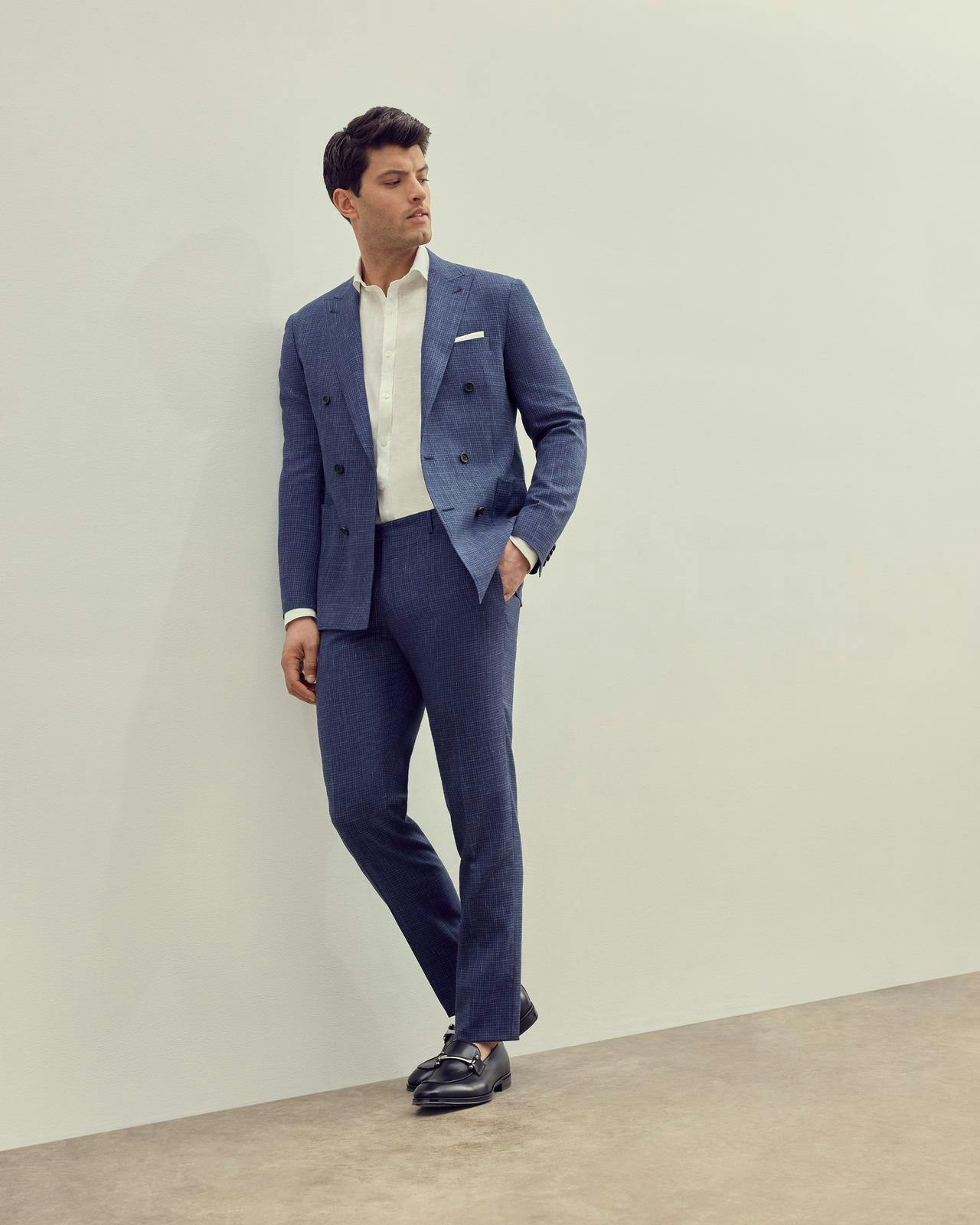 a male model in a blue suit and white t-shirt leaning against a wall