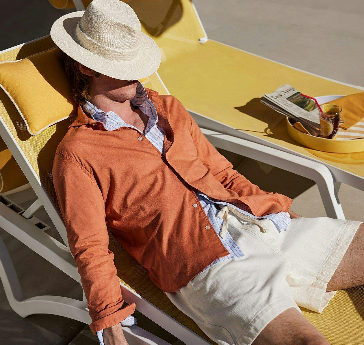 male model laying down on chair outside wearing sport shirt and shorts and hat covering face