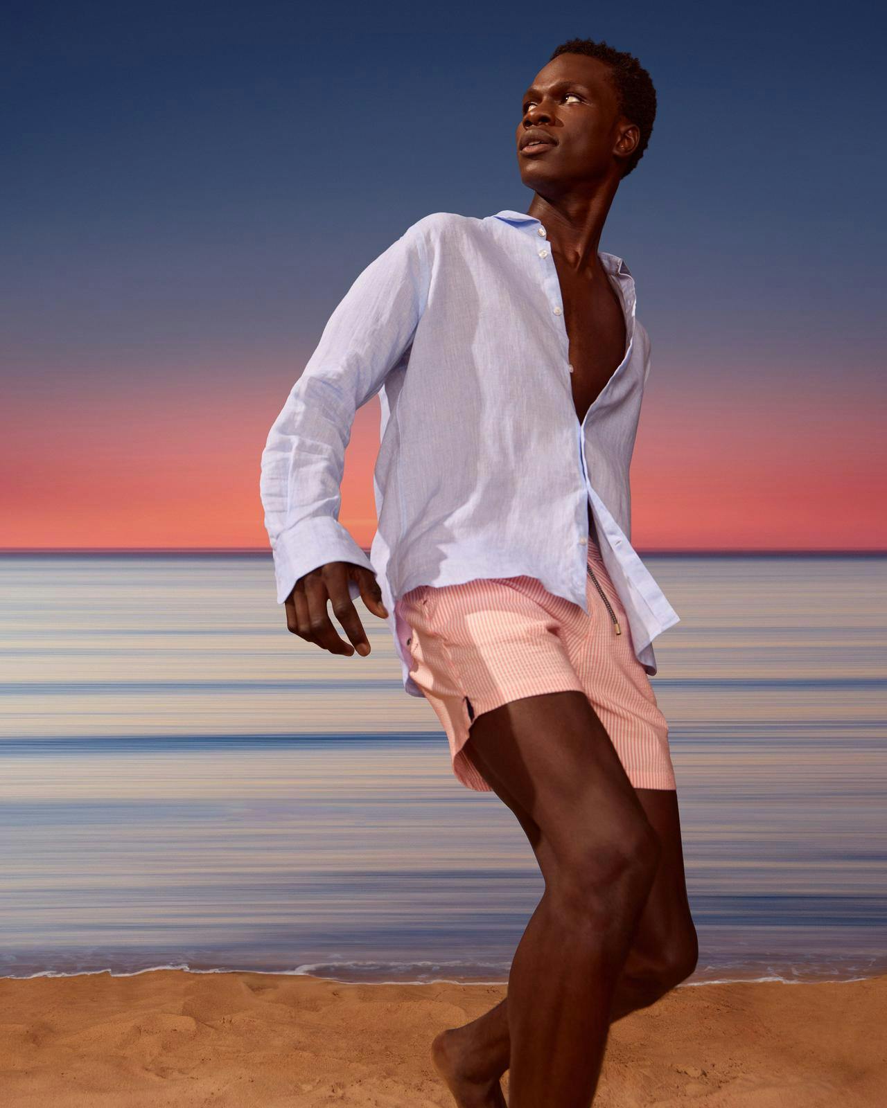 Un homme en chemise blanche et en shorts roses debout sur une plage.
