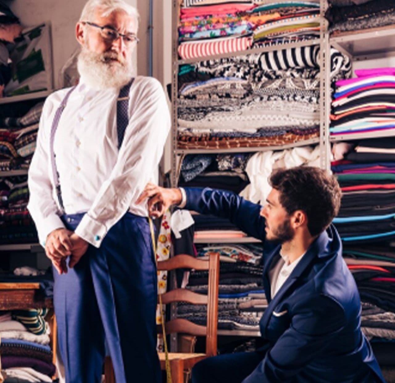 fabric backdrop with gentleman standing infront of chair wearing dress shirt and dress pants getting measured by gentleman