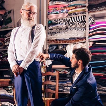 fabric backdrop with gentleman standing infront of chair wearing dress shirt and dress pants getting measured by gentleman