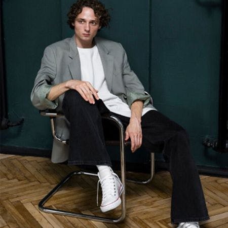 modèle masculin assis sur une chaise avec la jambe sur la poignée et la main sur le genou, porter une veste de sport, un t-shirt, un pantalon et des baskets