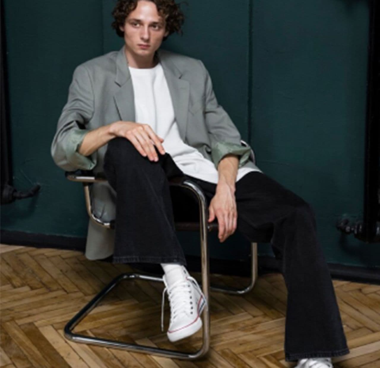 male model sitting on chair with leg over handle and hand on knee, wearing sport jacket, t-shirt, pants and sneakers