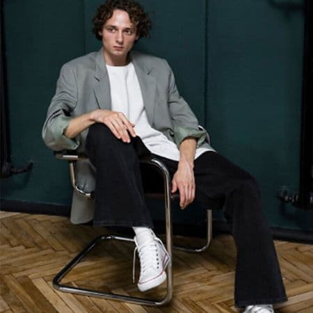 male model sitting on chair with leg over handle and hand on knee, wearing sport jacket, t-shirt, pants and sneakers