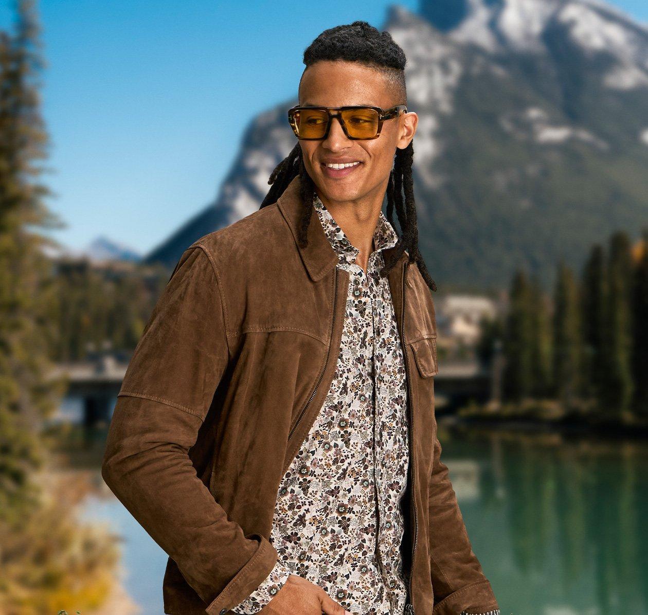 male model wearing sunglasses, dress shirt, jeans and overshirt outside infront of mountain scenery