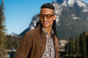 male model wearing sunglasses, dress shirt, jeans and overshirt outside infront of mountain scenery