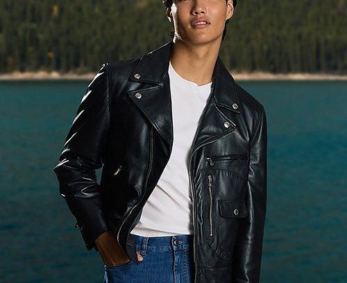 male model infront of lake and trees with one hand in pocket wearing leather jacket t-shirt and jeans