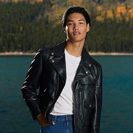 male model infront of lake and trees with one hand in pocket wearing leather jacket t-shirt and jeans