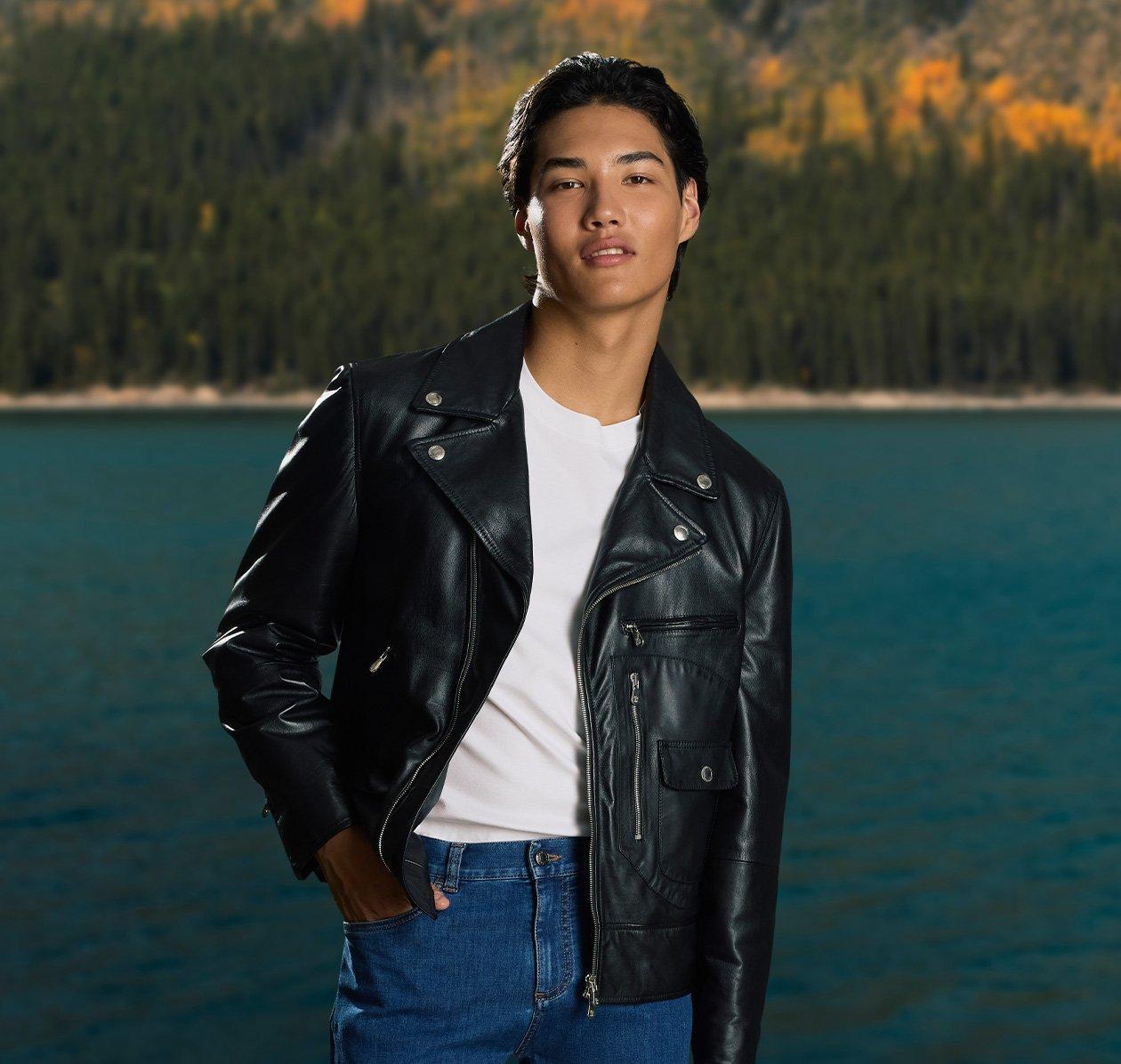 male model infront of lake and trees with one hand in pocket wearing leather jacket t-shirt and jeans