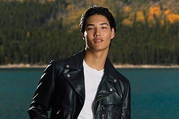 male model infront of lake and trees with one hand in pocket wearing leather jacket t-shirt and jeans